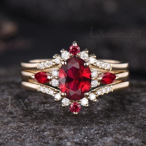 Puede incluir: Un conjunto de anillo de compromiso de tres piezas de rubí y diamantes en oro amarillo. La piedra central es un gran rubí ovalado rodeado por un halo de diamantes más pequeños. La banda está adornada con rubíes y diamantes más pequeños.