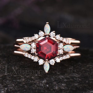 Puede incluir: Un juego de anillos de compromiso de tres piezas en oro rosa con una piedra preciosa roja hexagonal rodeada de piedras preciosas blancas más pequeñas. El anillo también tiene dos piedras preciosas blancas más pequeñas a cada lado de la piedra central.
