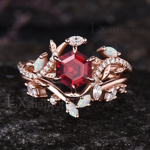 Può includere: Un anello di fidanzamento in oro rosa con una pietra rossa esagonale circondata da pietre bianche più piccole. L'anello presenta un delicato design floreale con foglie e rami.