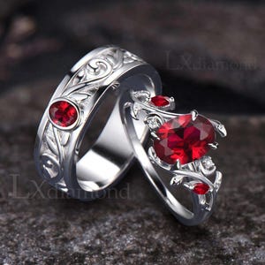 May include: Two silver rings with red gemstones. One ring has a central oval-cut ruby with smaller rubies and diamonds. The other ring has a round ruby and a floral design. The rings are set against a dark background.