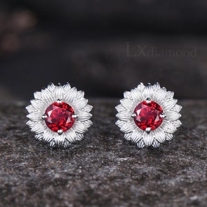 May include: A pair of silver stud earrings with a red gemstone in the center of each earring. The earrings are shaped like a flower with petals around the gemstone.