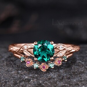 May include: A rose gold ring set featuring a round-cut dark green center stone flanked by leaf-shaped accents with small white stones. The matching curved band displays a series of rose-shaped metal details set with tiny green and pink stones.