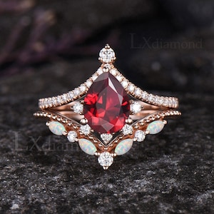 May include: A pear-shaped red gemstone engagement ring set with a matching band featuring white opal accents and a delicate diamond halo. The ring is set in rose gold.