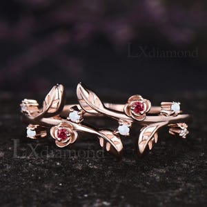 May include: Rose gold ring with a floral design. The ring features leaves, small red gemstones, and white gemstones. The ring is on a dark surface. The text "Lxdiamond" is visible.