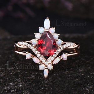 Puede incluir: Un anillo de piedra roja en forma de pera con un halo de ópalos blancos y piedras blancas más pequeñas. El anillo está engastado en oro rosa y tiene una banda a juego con un diseño delicado.
