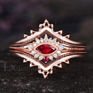 Könnte beinhalten: Roségoldener Ring mit einem zentralen, oval geschliffenen Rubin, umgeben von kleinen Diamanten. Der Ring hat ein einzigartiges, verziertes Design mit zusätzlichen kleinen Edelsteinen und einem geschichteten Band. Auf dunklem Hintergrund.