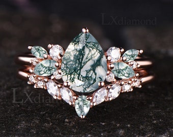 May include: A rose gold engagement ring with a pear-shaped green moss agate gemstone surrounded by smaller clear gemstones. The ring is set on a band with a delicate floral design.
