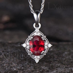 May include: A silver chain necklace with a pendant featuring a large oval-shaped red gemstone surrounded by smaller white gemstones in a delicate, ornate setting.