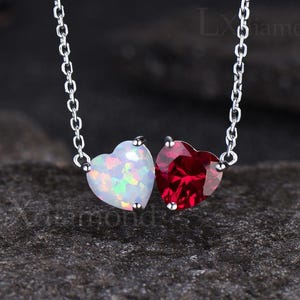 May include: A silver chain necklace with two heart-shaped pendants. The pendant on the left is white opal and the pendant on the right is red.