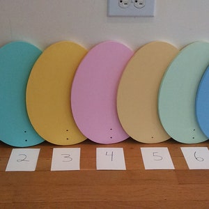 K&ouml;nnte beinhalten: Sieben ovale, aus Holz gefertigte Eier, die in Pastellfarben bemalt sind: lila, t&uuml;rkis, gelb, rosa, beige, hellgr&uuml;n und blau. Die Eier sind von 1 bis 7 nummeriert.