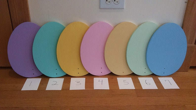 K&ouml;nnte beinhalten: Sieben ovale, aus Holz gefertigte Eier, die in Pastellfarben bemalt sind: lila, t&uuml;rkis, gelb, rosa, beige, hellgr&uuml;n und blau. Die Eier sind von 1 bis 7 nummeriert.
