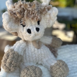 May include: A handmade crochet cow plushie in cream and brown. The cow has a fluffy, textured body with brown accents on the hooves, ears, and a patch on the chest. The eyes are black, and the horns are a lighter brown. The plushie is sitting on a white, textured surface.