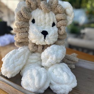 May include: A handmade plush lion toy with a cream-colored body and a tan mane. The lion has black embroidered eyes and a black nose. The toy is made of soft, chunky yarn and is sitting on a wooden tray.