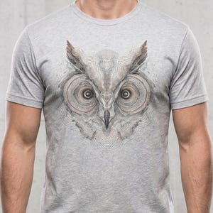 May include: A heather grey short-sleeve t-shirt featuring a detailed graphic print of an owl face on the chest. The owl illustration is rendered in shades of brown, black, and white with intricate line work that mimics topographic map contours.