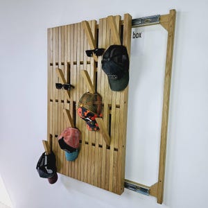 May include: Wooden wall-mounted coat rack with multiple hooks, holding sunglasses and various baseball caps. The rack is made of light brown wood and has a sliding door mechanism. The word "box" is visible on the right side.