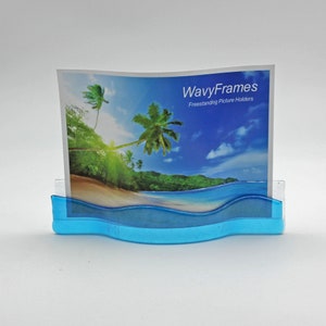 May include: A blue plastic picture frame with a wavy design. The frame is holding a photo of a tropical beach scene with palm trees and blue water. The text "WavyFrames Freestanding Picture Holders" is printed on the photo.