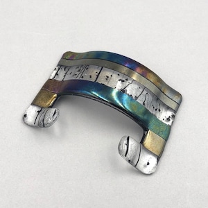 May include: A silver and gold metal brooch with a wavy, abstract design. The brooch has a iridescent finish with blue, green, and purple hues.