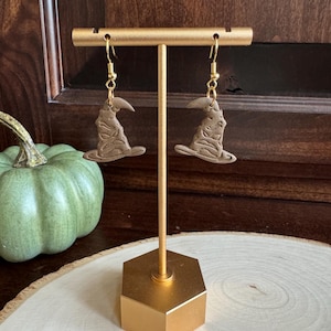 May include: A pair of brown, ceramic earrings shaped like the Sorting Hat from the Harry Potter series. The earrings are hanging from gold hooks.