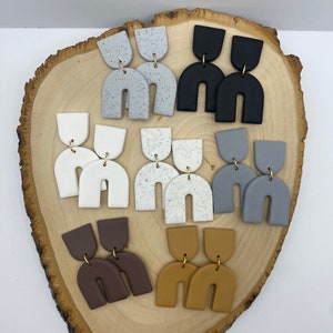 Puede incluir: Surtido de 10 pares de pendientes hechos a mano de arcilla polimérica. Los pendientes son de varios colores, incluyendo blanco, gris, marrón, negro y tostado. Cada par presenta una forma de arco única con un pequeño acento dorado.