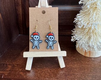 Puede incluir: Un par de pendientes colgantes con un esqueleto negro que lleva un gorro de Papá Noel rojo. Los pendientes se muestran en un pequeño caballete de madera.