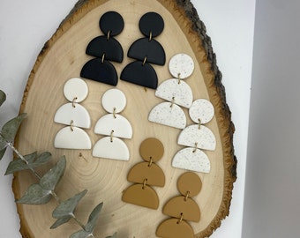 May include: Three pairs of statement earrings displayed on a wooden surface. The earrings are made of polymer clay and feature a geometric design. The first pair is black, the second pair is white, and the third pair is brown.