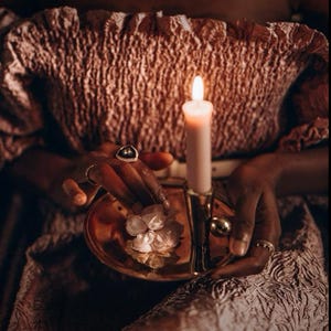 May include: A lit pink candle in a gold-colored holder. The candle is held over a small, round, gold-colored dish with a white flower. The person is wearing rings on their fingers and a pink dress.