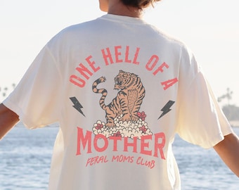 May include: A white t-shirt with a graphic of a tiger and the text "One Hell of a Mother Feral Moms Club".