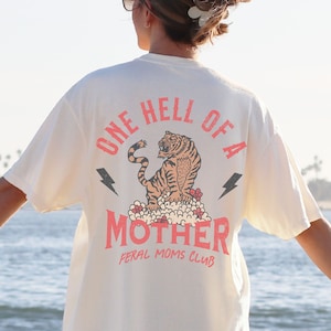 May include: A white t-shirt with a graphic of a tiger and the text "One Hell of a Mother Feral Moms Club".