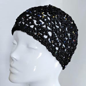 May include: A black crocheted hat with a net-like design, adorned with colorful sequins. The hat is displayed on a white mannequin head, showcasing its texture and style. The sequins add a touch of sparkle to the dark, open weave.