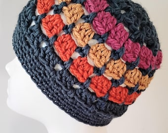 May include: A crocheted beanie featuring a dark charcoal gray base with a band of alternating square clusters in shades of burnt orange, tan, and magenta. The hat is constructed with an openwork stitch pattern and a solid dark gray brim.