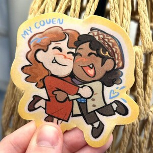 May include: A die-cut sticker featuring a cartoon illustration of two characters hugging. One character has wavy reddish-brown hair and a red coat, while the other has dark curly hair, a patterned hat, and a beige coat. The text MY COVEN is printed.