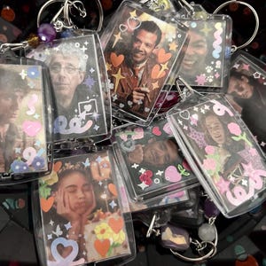 Puede incluir: Una colección de llaveros rectangulares transparentes con varias imágenes de personas. Cada llavero tiene un borde decorativo con corazones, estrellas y elementos florales. Algunos llaveros tienen detalles de cuentas.