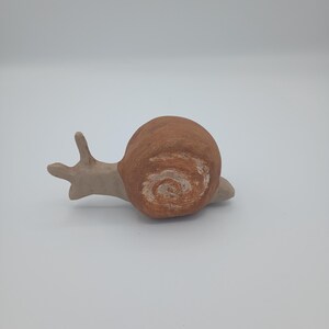 May include: A handmade ceramic snail figurine. The snail has a brown shell with a white spiral design and a light tan body. The snail's antennae are extended, and it appears to be crawling.
