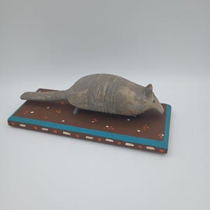 May include: A hand-painted wooden armadillo figurine on a brown base with white and orange accents. The armadillo is gray and has a textured surface.