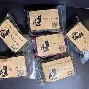 May include: Assortment of dried herbs and botanicals in clear plastic bags. Each bag has a label with the text "Black Cat Botanica" and the name of the herb, such as "Basil Leaf" or "Rose Petals". A black cat illustration is on each label.