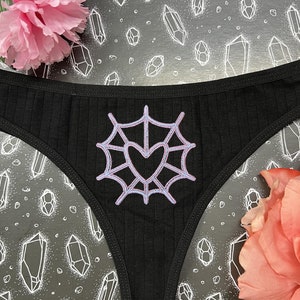 Könnte beinhalten: Schwarze Spitzenunterwäsche mit einem weißen Spinnennetz und einem rosafarbenen Herzdesign.
