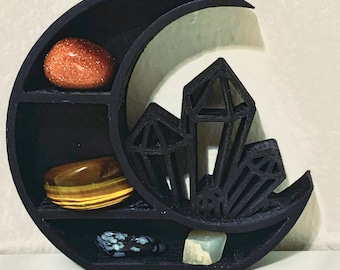 May include: Black crescent moon-shaped shelf with five compartments for displaying crystals and other small objects. The shelf is decorated with a crystal design.