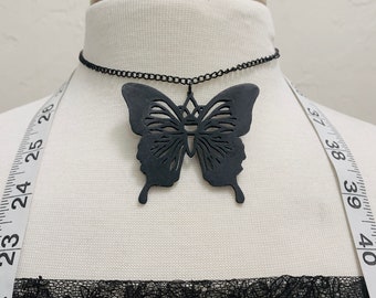 Puede incluir: Un collar gargantilla negro con un gran colgante de mariposa intrincado. La mariposa está recortada y tiene un diseño geométrico en el centro.