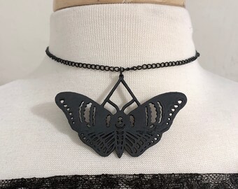 Puede incluir: Un collar gargantilla negro con un gran colgante de mariposa cortado de forma intrincada. La mariposa está hecha de un material negro y tiene un diseño delicado y de encaje.