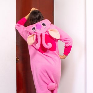 Puede incluir: Pijamas de una pieza rosa para adulto con una gran cabeza de elefante rosa en la capucha. El pijama tiene una cola rosa y detalles rosas en los pu&ntilde;os y los tobillos.