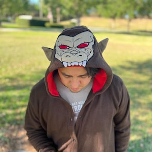 May include: A brown hooded jumpsuit with a red lining and a large embroidered monkey head on the hood. The monkey head has red eyes and sharp teeth. The person wearing the jumpsuit is looking down at a black smartphone.