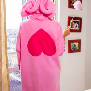 Puede incluir: Un mono rosa con capucha y un gran coraz&oacute;n en la espalda. El mono tiene una cola y orejas peludas. La persona que lleva el mono est&aacute; de pie frente a una ventana y sostiene un plumero.