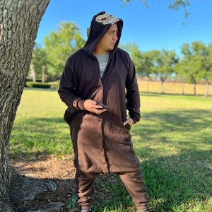 May include: A brown hooded jumpsuit with a cartoon animal face on the hood. The jumpsuit has a zip and buttons down the front. The person wearing the jumpsuit is standing in a grassy area next to a tree.