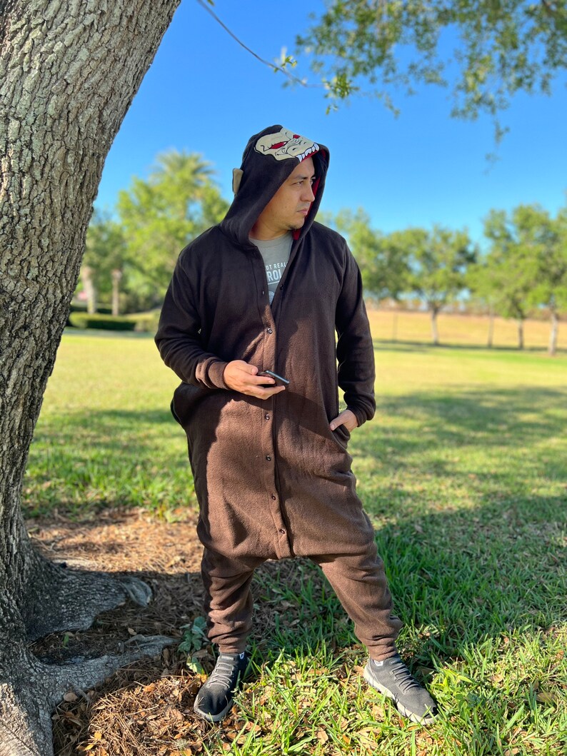 May include: A brown hooded jumpsuit with a cartoon animal face on the hood. The jumpsuit has a zip and buttons down the front. The person wearing the jumpsuit is standing in a grassy area next to a tree.