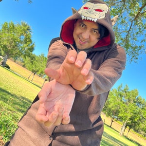 May include: A person wearing a brown hooded jumpsuit with a red and white monster face on the hood. The person is smiling and has their hands up in a claw-like gesture.