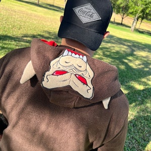 May include: A brown hooded sweatshirt with a cartoon monster face on the hood. The person is wearing a black baseball cap with a grey diamond-shaped patch that says "Drink Coca-Cola in Bottles".