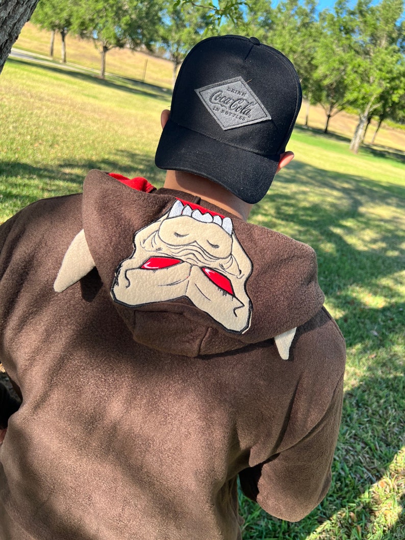 May include: A brown hooded sweatshirt with a cartoon monster face on the hood. The person is wearing a black baseball cap with a grey diamond-shaped patch that says "Drink Coca-Cola in Bottles".