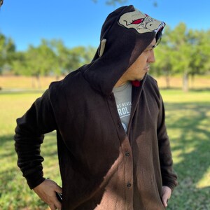 May include: A brown fleece hooded cardigan with a button fastening. The hood features a large embroidered design of a creature with red eyes. The text "Not Really" and "ROL" is visible on the front of the cardigan.