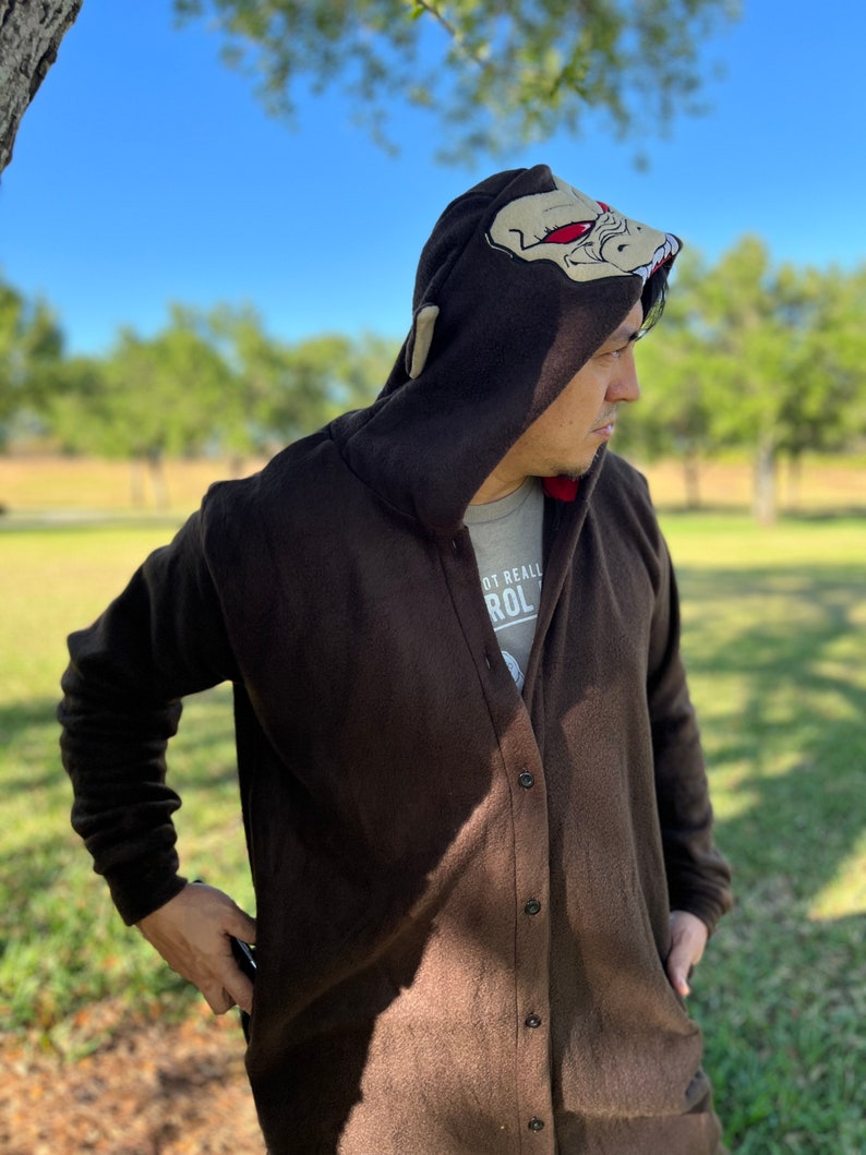 May include: A brown fleece hooded cardigan with a button fastening. The hood features a large embroidered design of a creature with red eyes. The text "Not Really" and "ROL" is visible on the front of the cardigan.