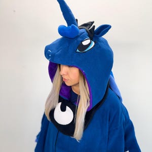 Peut inclure: Un vêtement à capuche en polaire bleu foncé avec une corne de licorne, des oreilles pointues et des yeux brodés. L'intérieur de la capuche est violet et une pièce de cou noire affiche un emblème de croissant de lune blanc avec fermeture éclair.